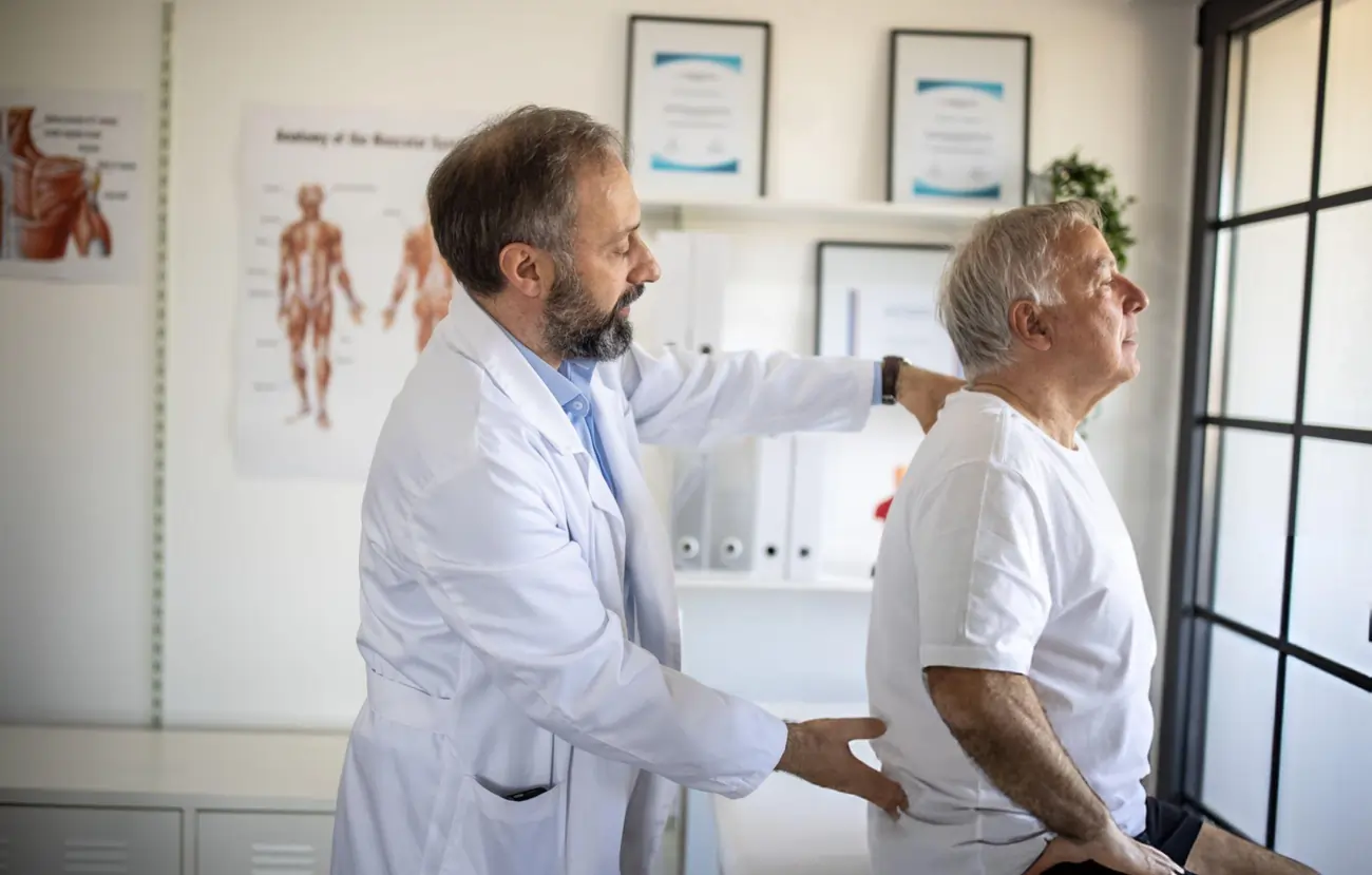 A male doctor in a white coat examines the lower back of an older male patient, representing a physical exam for back pain or a spine condition as part of a consultation for stem cell therapy in Battle Creek, Michigan.