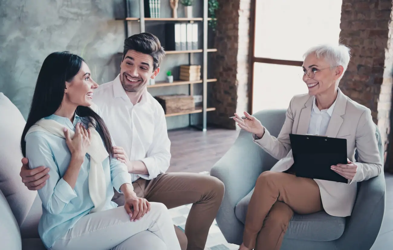 A smiling couple, a man and a woman, sitting on a couch and happily conversing with an older female doctor or consultant, representing a successful patient consultation for stem cell therapy in Battle Creek, Michigan.