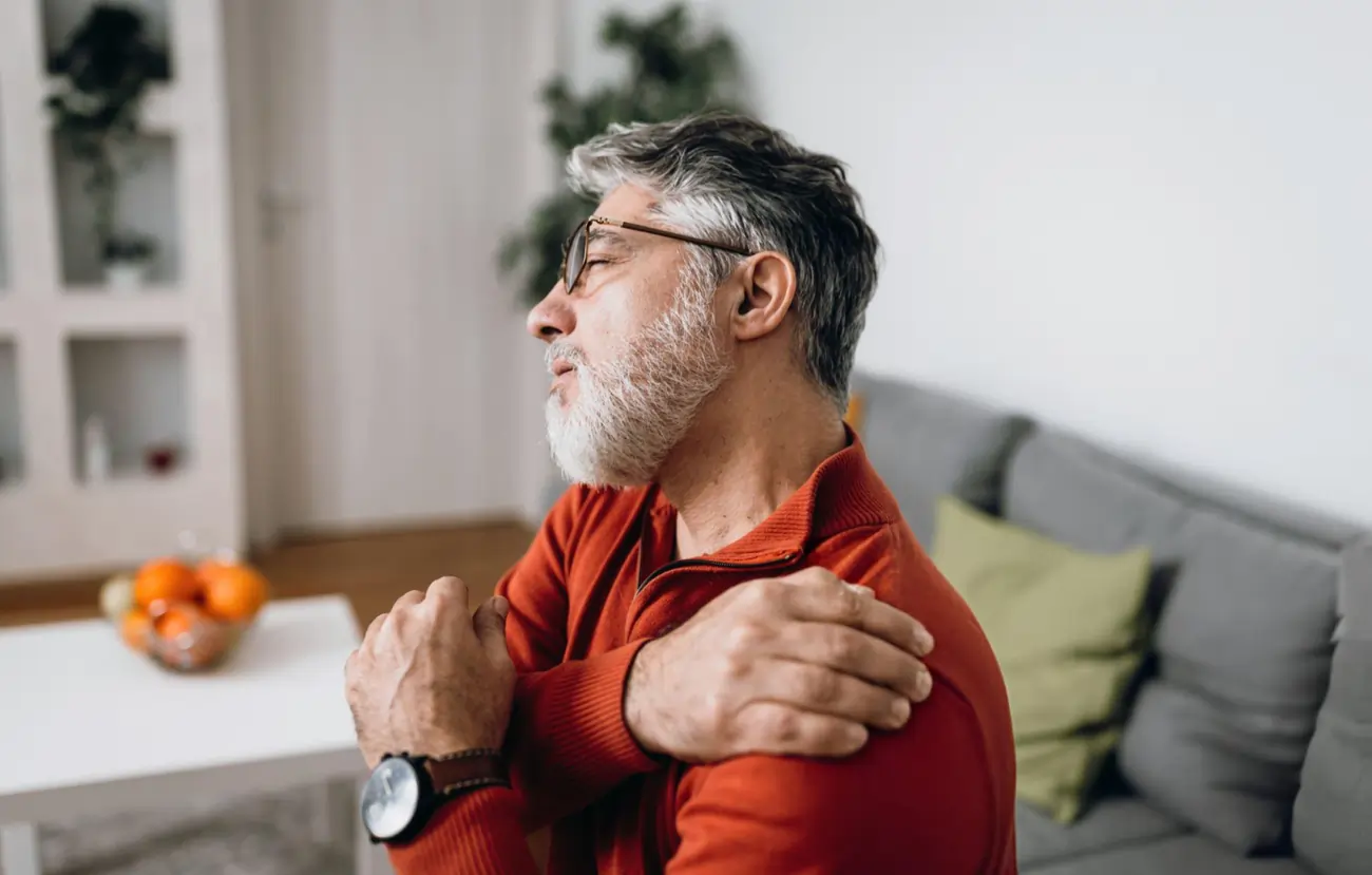 Middle-aged man with a gray beard and glasses, sitting on a couch and holding his shoulder in pain, representing a patient seeking stem cell therapy for joint or shoulder pain in Battle Creek, Michigan.