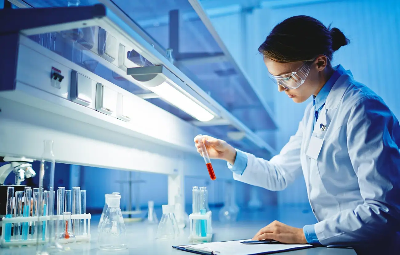 A dedicated female scientist or lab technician, wearing a lab coat and safety goggles, examines a blood sample in a test tube inside a modern laboratory.