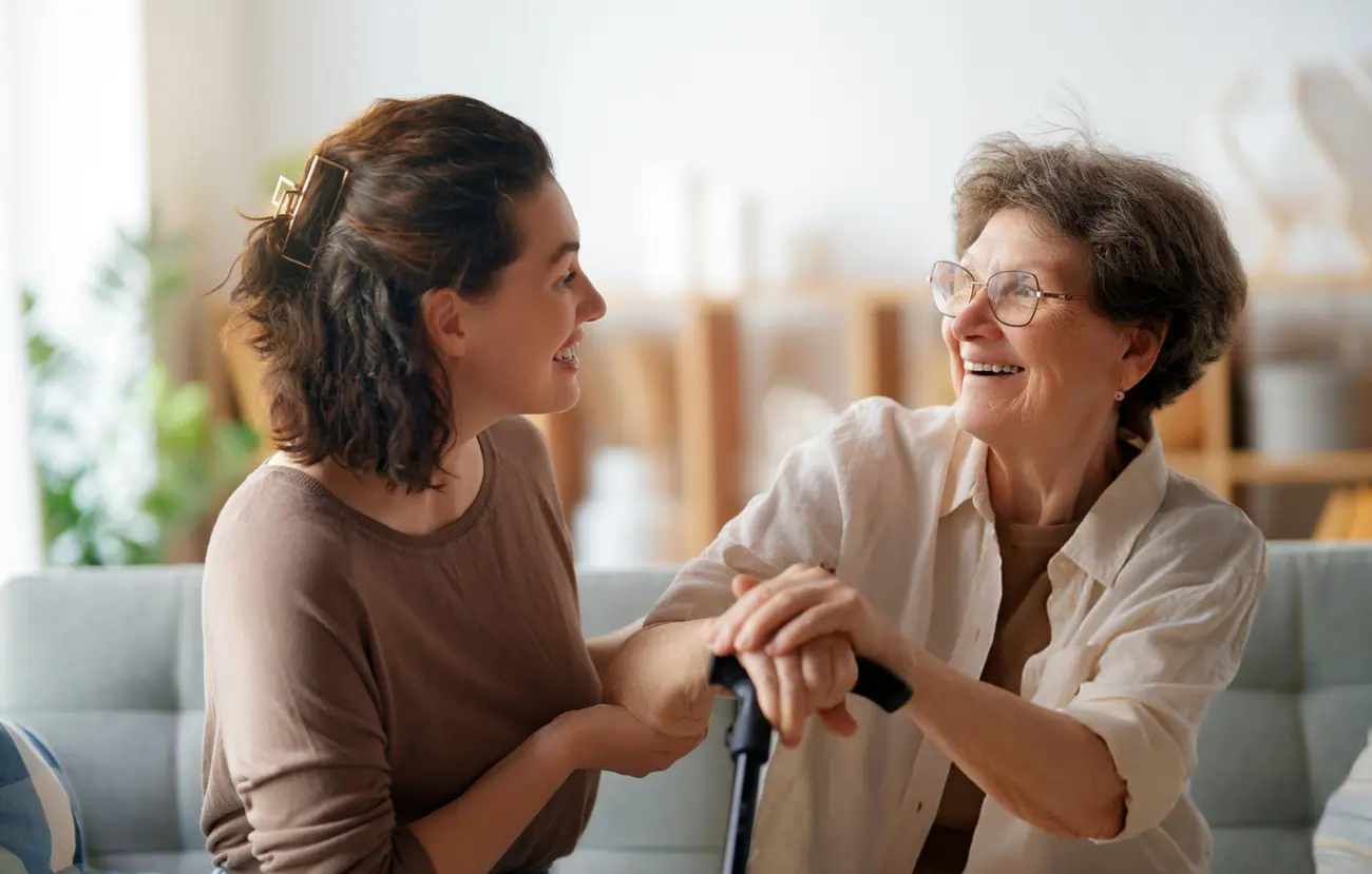 A kind caregiver assists a senior woman with a cane, both smiling and sharing a happy moment on a couch, representing compassionate care for elderly patients.